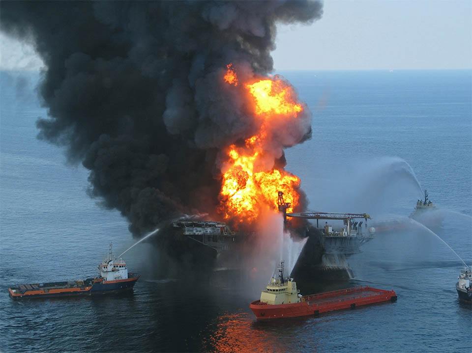 Luftaufnahme von einem Brand auf einer Bohrinsel. Zentral sind die lodernden Flammen und die dunklen, schwarzen Rauchschwaben. Im umgebenden Meer sind vier Löschfahrzeuge bzw. Schiffe, welche Wasserstrahlen ins Feuer leiten.