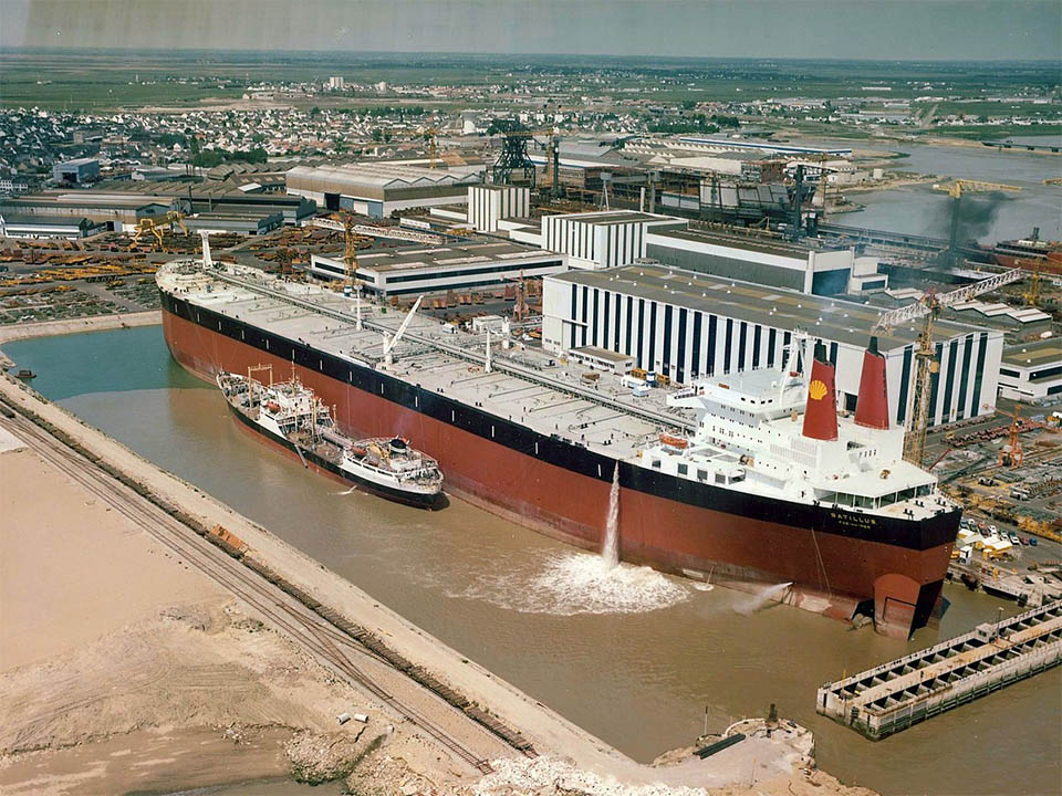 Abbild des Batillus Tankers in Saint-Nazaire. Der rießige rote Öltanker steht Mitten in dem Industriehafen und schwimmt auf trübem braunen Wasser. 