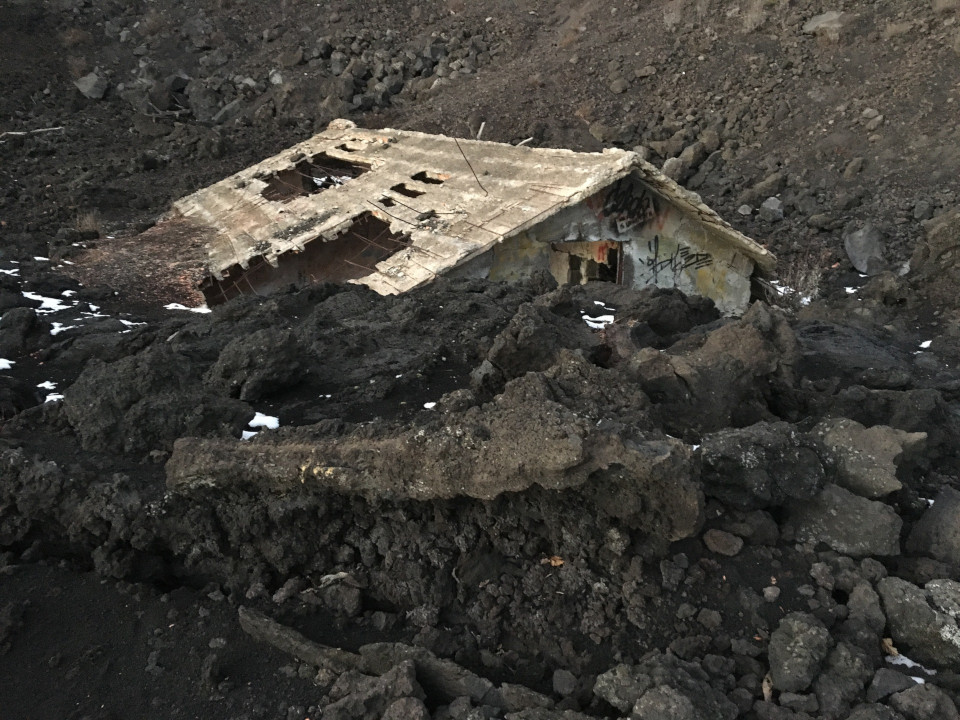 Ein Haus, dessen Dach sichtbar ist, der Rest wurde von Lava zerstört und liegt nun in schwarzem Vulkagestein begraben.